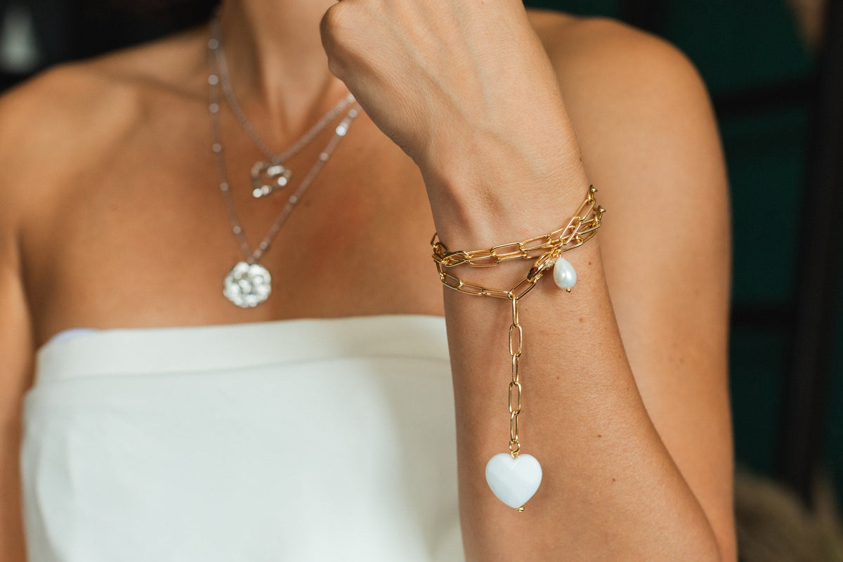 A woman's wrist in a gold chain bracelet with a white heart charm, in front of layered silver necklaces