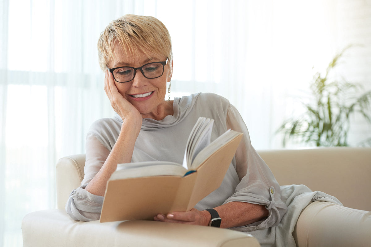 A woman in glasses relaxing on a sofa with a book