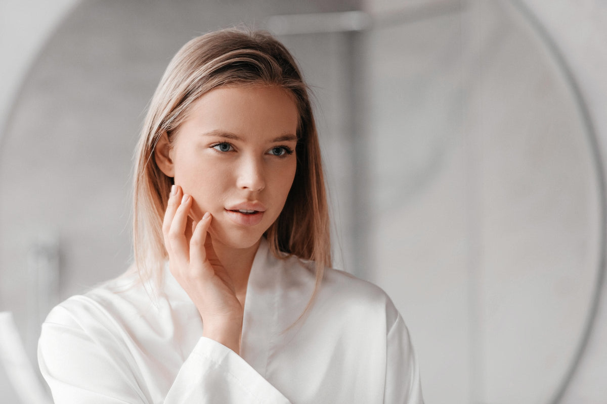 A woman in a white robe gently touching her cheek in front of a bathroom mirror