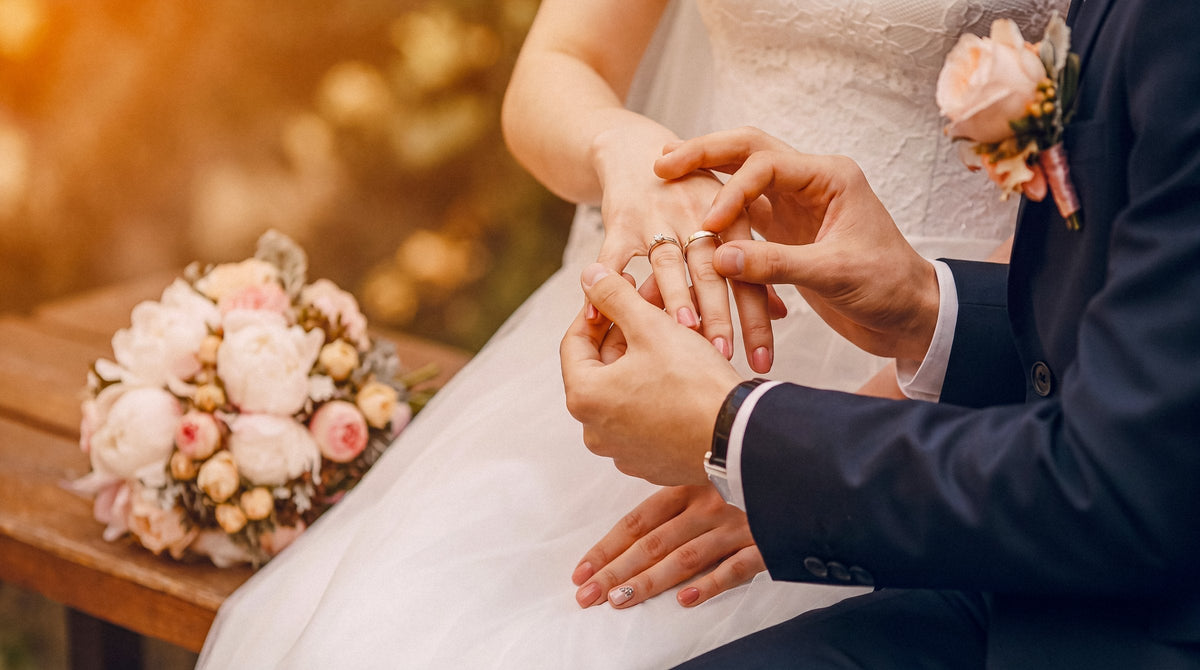 A wedding ring being placed on a bride's finger at an outdoor ceremony, a bouquet beside them
