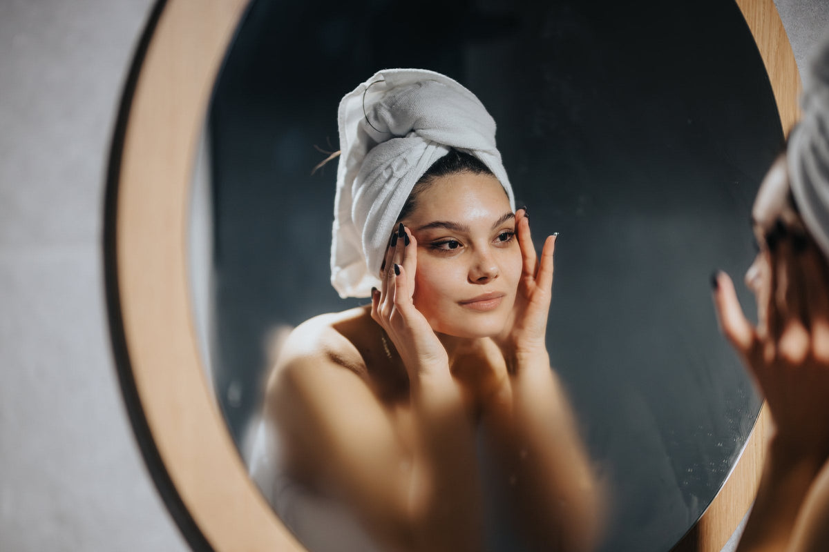 A woman in a towel touching her face at a round mirror in the evening